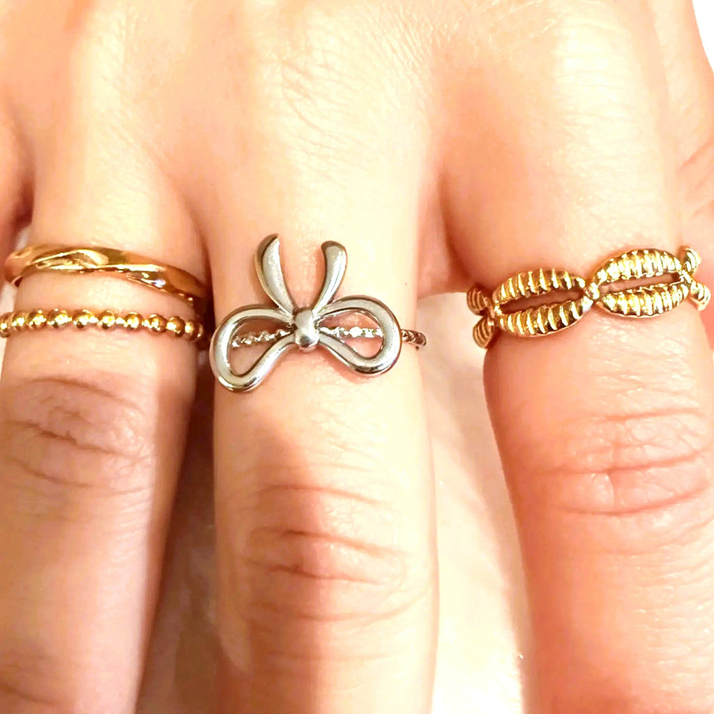 A close-up of a hand wearing three rings: a silver bow-shaped ring on the middle finger, a thin gold beaded band on the ring finger, and a textured gold wrap ring on the ring finger.