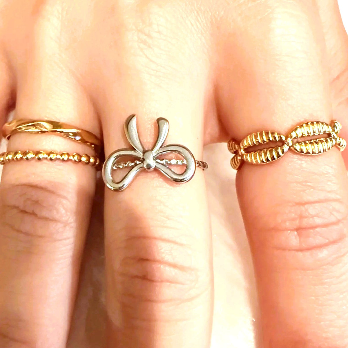 A close-up of a hand wearing three rings: a silver bow-shaped ring on the middle finger, a thin gold beaded band on the ring finger, and a textured gold wrap ring on the ring finger.